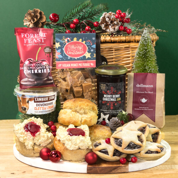 The Gluten Free Devon Christmas Cream Tea features scones with clotted cream and jam, mince pies, fudge, chocolate dipped cherries, and tea, all beautifully arranged with festive decorations on a wooden platter.