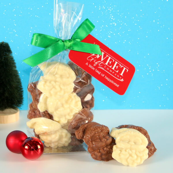 A clear bag tied with a green ribbon contains white and milk chocolate sheep-shaped candies. The bag has a red tag with "A little bag of happiness." In the background, small ornaments, a tree, and Milk & White Chocolate Santas can be seen.