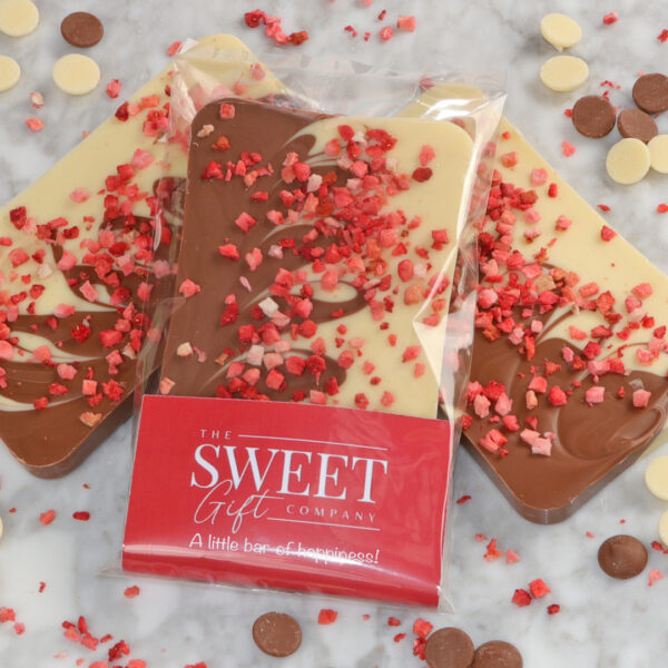 Three chocolate bars, including The Classic Strawberries & Cream Bar, featuring a blend of white and milk chocolate topped with strawberry pieces, are displayed on a marble surface. One bar is partially wrapped in branded packaging reading "The Sweet Gift Company, "A little bar of happiness!"