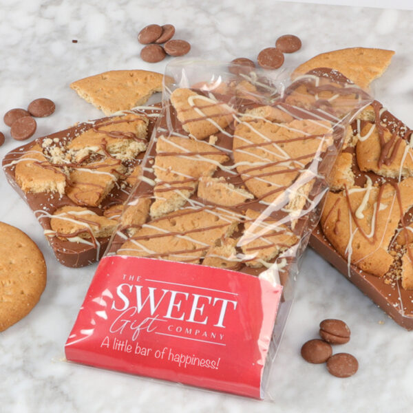 Three milk chocolate bars, including The Delicious Digestive Bar, topped with digestive biscuits and milk chocolate drizzle, are displayed on a marble surface. One bar is partially wrapped in branded packaging reading "The Sweet Gift Company, "A little bar of happiness!"