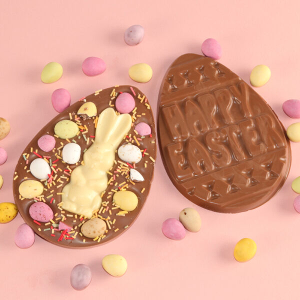 Chocolate Easter egg on a pink background: one with a white chocolate bunny and candy decorations, and the other is showing the back of The Mini Egg Chocolate Easter Egg Slab elegantly embossed with "Happy Easter.
