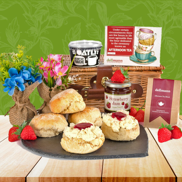 A slate platter with scones, clotted cream, jam, and strawberries from The Vegan Cream Tea Hamper sits before a wicker hamper, tea, and decor on a wooden table set against a green patterned backdrop.
