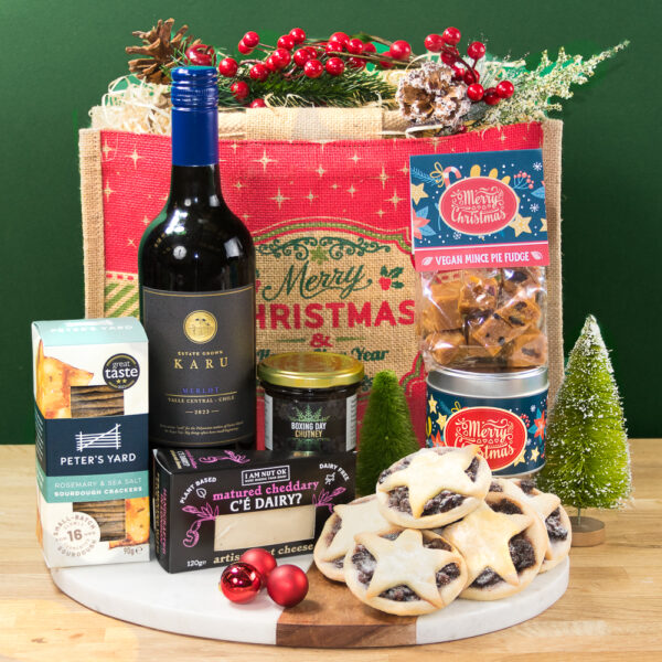 The Vegan Christmas Yuletide Hamper includes wine, vegan cheese, a gluten-free Christmas cracker, mince pies, fudge, chutney, and festive decor on a wooden board with a red "Merry Christmas" gift bag in the background.